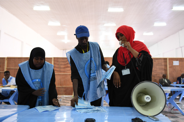 Eine Gruppe von Menschen steht um einen Tisch mit einem Megafon, Papieren und anderen Gegenständen herum, die wie eine Wahllokale aussehen, während andere im Hintergrund sitzen und stehen.