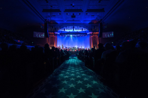 Ein großer Zuschauerraum mit Menschen auf Stühlen, die einer Bühne mit Musikern zugewandt sind, unter einer Decke mit Lampen und Fernsehgeräten, was auf eine Konzerthalle hinweist.