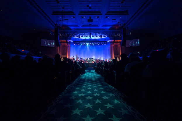 Ein großer Zuschauerraum mit Menschen auf Stühlen, die einer Bühne mit Musikern zugewandt sind, unter einer Decke mit Lampen und Fernsehgeräten, was auf eine Konzerthalle hinweist.