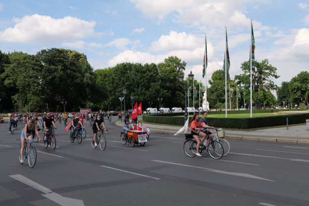 Eine Gruppe von Menschen, die auf Fahrrädern eine von Bäumen gesäumte Straße während der Paris Bike Ride 2016 entlangfahren, mit Laternenmasten, Fahnenmasten, bewölktem Himmel und Fahrzeugen im Hintergrund.