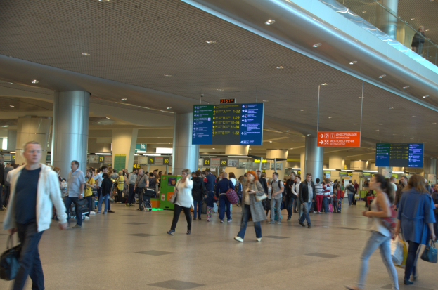 Eine große Gruppe von Menschen geht durch einen Flughafen, einige tragen Gepäck, über Kopf Schilder, Deckenleuchten, Säulen und ein Glasgeländer auf der rechten Seite.
