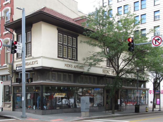 Eine belebte Stadtstraße mit einem geparkten Bus, Gebäuden, Bäumen, Laternen, Verkehrszeichen, Schildern und einem Kaufhaus mit Schaufensterpuppen in Kleidern vor einem sichtbaren Himmel.