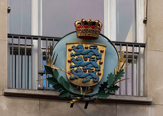 Wappen der britischen Königsfamilie an einer Gebäudewand mit einer Krone und einem Blatt-Logo, Fenstern und Gittern im Hintergrund.