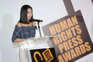 Eine Frau in einem schwarzen Kleid steht an einem Rednerpult mit einem Mikrofon und hält eine Rede oder Präsentation vor einem Banner, auf dem "Human Rights Press Awards" steht, ihr Haar ist zurückgebunden und ihr Gesichtsausdruck ernst und konzentriert.