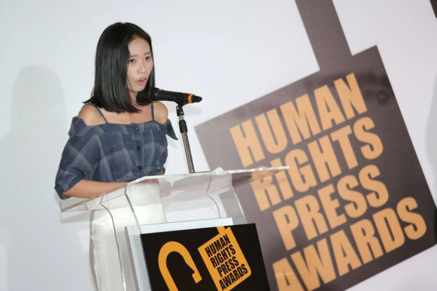 Eine Frau in einem schwarzen Kleid steht an einem Rednerpult mit einem Mikrofon und hält eine Rede oder Präsentation vor einem Banner, auf dem "Human Rights Press Awards" steht, ihr Haar ist zurückgebunden und ihr Gesichtsausdruck ernst und konzentriert.
