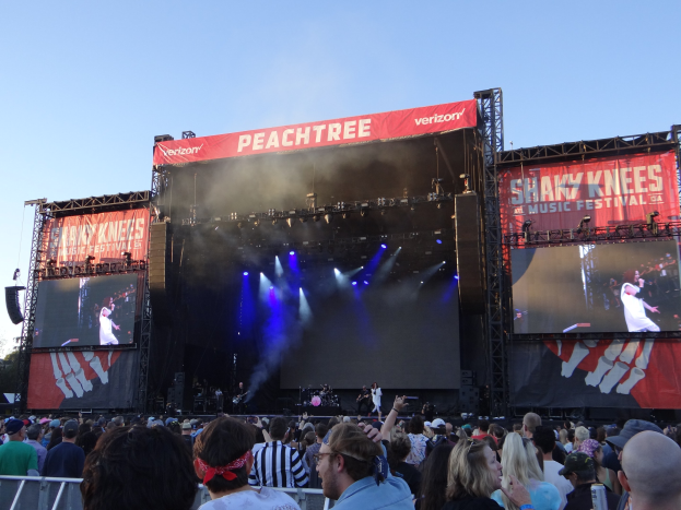 Eine große Menschenmenge steht vor einer hell erleuchteten Bühne beim Shaky Knees Music Festival, auf der Musiker mit Instrumenten und Schildern hinter einem Geländer unter einem klaren blauen Himmel spielen.