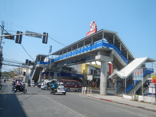 Eine belebte Stadtstraße mit Fahrzeugen, Fußgängern, Verkehrsampeln, Gebäuden, einer Mautstelle und einem klaren blauen Himmel mit Bäumen im Hintergrund.