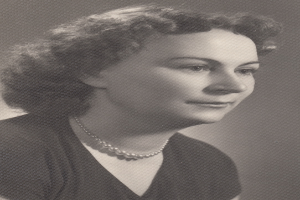 Ein Schwarz-Weiß-Foto einer Frau mit ernstem Gesichtsausdruck, deren Haar zurückgekämmt ist und die eine Kette sowie ein einfaches Kleid mit hohem Kragen und langen Ärmeln trägt.