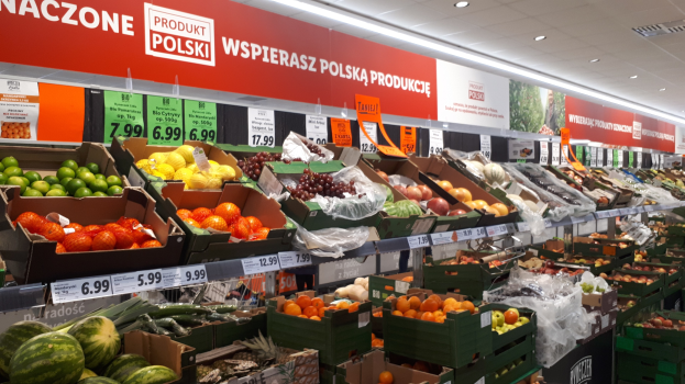 Ein Geschäft mit Kartons voller verschiedener Obst- und Gemüsesorten, jeweils mit Preisschildern, beleuchtet von Deckenlampen und mit Informationsschildern an den Wänden.