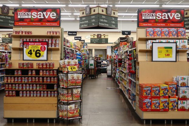 Ein Kiosk-Innenraum mit Regalen und Boden voller verschiedener Lebensmittel, Deckenbeleuchtung und Informationsschilder im Hintergrund.