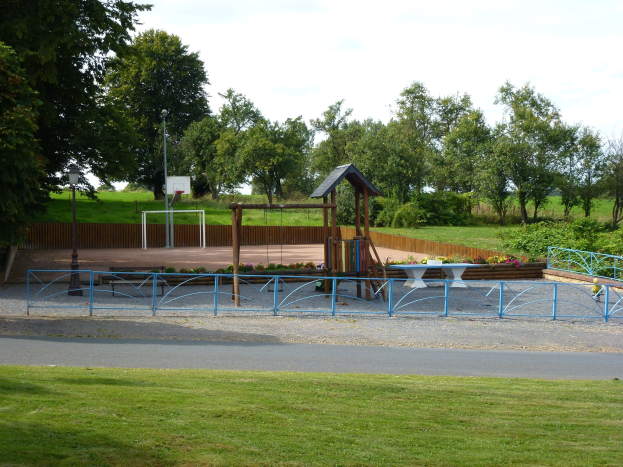 Ein Park mit einem Spielplatz in der Mitte, umgeben von einer Straße, Gras, Pflanzen, Blumen, einem Zaun, Pfosten, einem Basketball-Korb, Bäumen und einem klaren blauen Himmel.