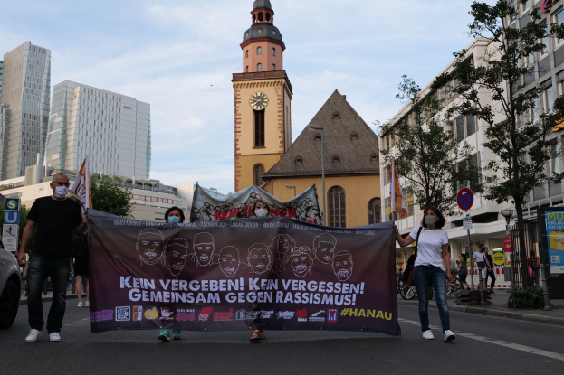 Eine Gruppe von Menschen mit Masken, die eine Straße entlanggehen und ein Banner halten, mit einem geparkten Auto auf der linken Seite, Gebäuden, Bäumen, Schildern, Pfählen, einem Uhrenturm und einem klaren blauen Himmel im Hintergrund.