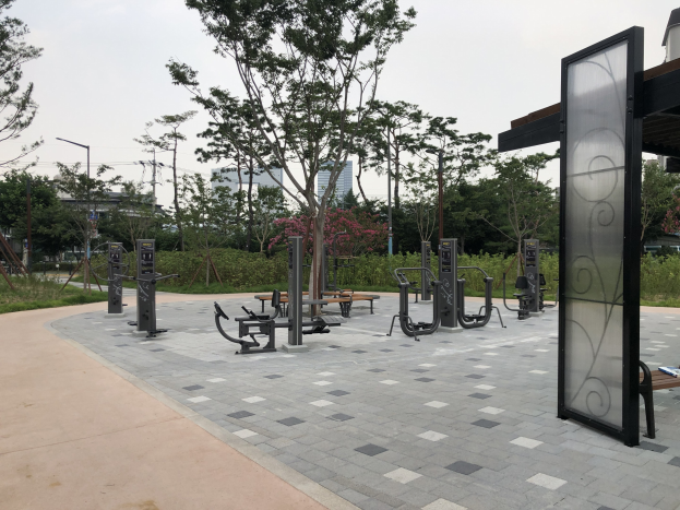 Ein Park mit Außen-Fitnessgeräten, Bänken, Bäumen, Pflanzen, Gras, Pfosten, Lichter, Drähte und Gebäude unter einem Himmel-Hintergrund.