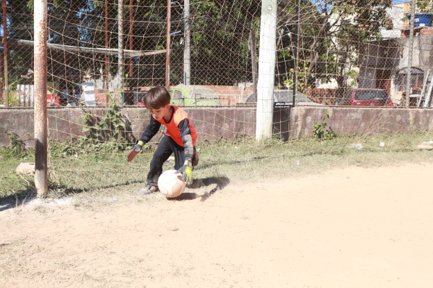 Ein junger Junge spielt Fußball auf einem staubigen Feld mit Gras, Pflanzen, Pfählen, einem Zaun, einer Wand, Bäumen, Fahrzeugen, Gebäuden und einem bewölkten Himmel im Hintergrund, trägt Schuhe und ist aktiv im Spiel.