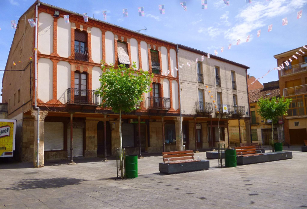 Ein Platz mit Gebäuden, Fenstern, Geländern, Bäumen, Bänken, Mülltonnen, Pfählen, Drähten und dekorativen Flaggen unter einem klaren blauen Himmel.