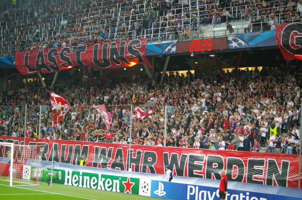 Ein Fußballspiel in einem Stadion mit einer großen Zuschauermenge, einem saftig grünen Rasen, einem Torpfosten links, Bodenbannern, Menschen mit Fahnen und Hintergrundgeländern, Tafeln, Lampen und anderen Gegenständen.