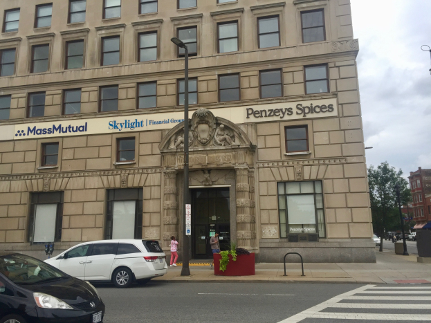 Großes Gebäude mit "Massachusetts Mutual Financial Group" -Schriftzug, parkende Autos davor, Straßenlaternen, fahrende Autos, Fußgänger auf dem Gehweg, Bäume und bewölkter Himmel.