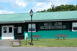 Ein Rathaus-Gebäude in Germantown, Pennsylvania, mit Fenstern, Türen und einem Dach, umgeben von Bänken, Gras, einem Straßenmast, einem Schild, einem Müllcontainer, Topfpflanzen mit Blumen, einer Baumgruppe und einem bewölkten Himmel.