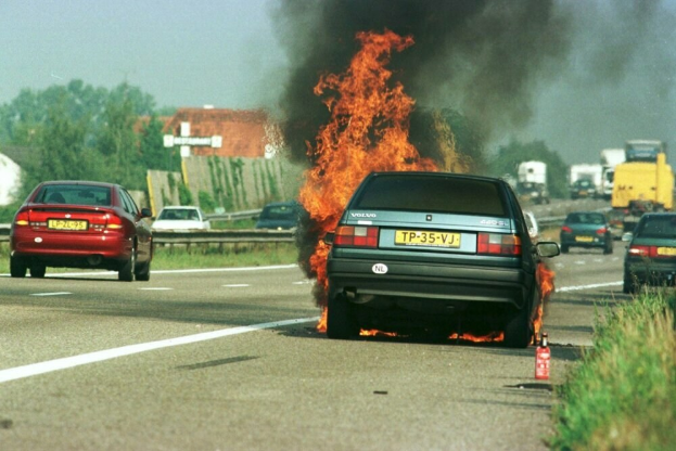 Ein Auto, das in Flammen steht, am Straßenrand mit anderen Fahrzeugen in der Nähe, Bäumen, Gebäuden und einem klaren blauen Himmel im Hintergrund und einem Feuerlöscher auf der rechten Seite.