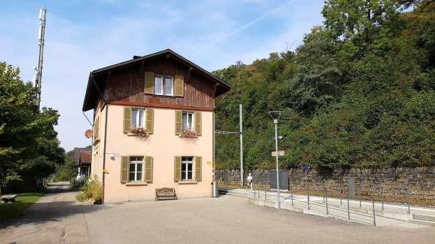Ferienhaus zum Verkauf in Baden-Württemberg, Deutschland, mit einem Gebäude mit zahlreichen Fenstern, einem vorderen Zaun, umgebender Straßenmöblierung, Grünfläche und einem bewölkten Himmel.