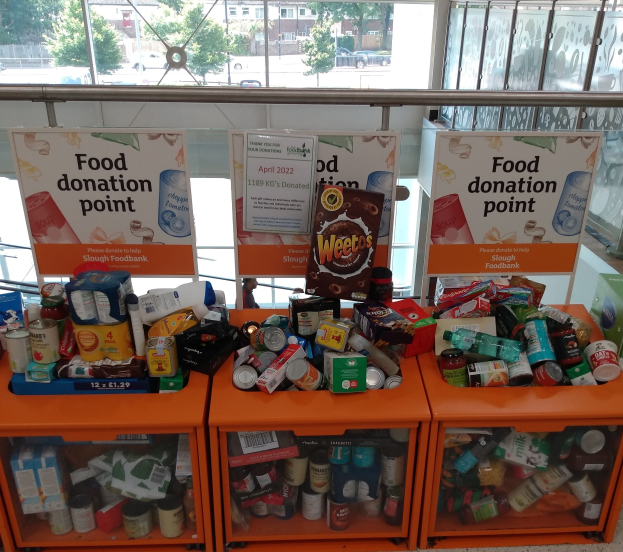 Orange Schränke in einem Gebäude mit Lebensmittelspenden wie Flaschen, Dosen und Kartons, mit Informationsschildern in der Nähe, durch Glasfenster betrachtet, die Outdoor-Fahrzeuge, Bäume, Gebäude und Himmel zeigen.