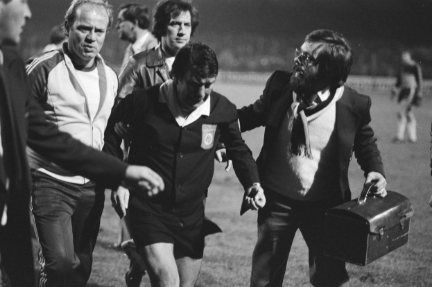 Schwarzes und weißes Bild einer Gruppe von Männern, die auf einem Fußballfeld gehen, einer hält eine Aktentasche, mit einem Fußballspieler in der Mitte, der besorgt aussieht.