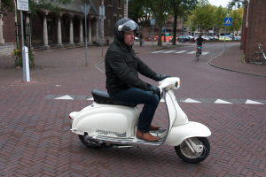 Ein Mann auf einem weißen Roller, der eine schwarze Jacke, blaue Jeans und einen schwarzen Helm trägt, fährt auf einer Straße mit anderen Menschen auf Fahrr√°dern, Autos, grünen Bäumen und braunen und cremefarbenen Gebäuden im Hintergrund unter einem hellen weißen Himmel.