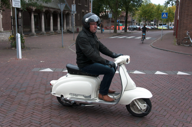 Ein Mann auf einem weißen Roller, der eine schwarze Jacke, blaue Jeans und einen schwarzen Helm trägt, fährt auf einer Straße mit anderen Menschen auf Fahrr√°dern, Autos, grünen Bäumen und braunen und cremefarbenen Gebäuden im Hintergrund unter einem hellen weißen Himmel.
