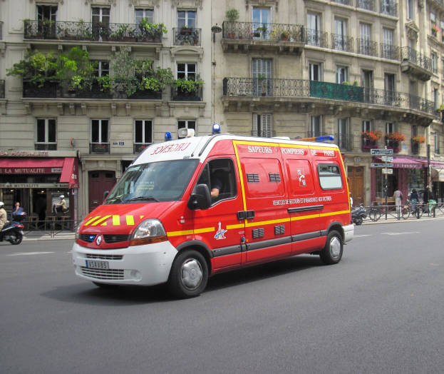Ambulanz auf der Seite der Straße vor einem Gebäude mit Fenstern, Geländern, Pflanzen und anderen Gegenständen geparkt, mit wenigen Menschen und Fahrrädern im Hintergrund.