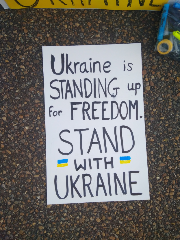 Ein weißes Blatt Papier mit dem Text "Ukraine steht für Freiheit ein, stellt euch mit Ukraine" vor einer gelben Fahne auf dem Boden. Rechts davon ein Plastikbehälter mit verschiedenen Gegenständen.