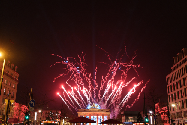 Eine belebte Straßen Szenerie in Berlin am Silvesterabend, voller Menschen, Fahrzeuge und festlicher Lichter von Gebäuden und Feuerwerk am Himmel.