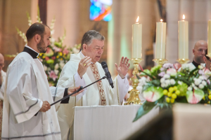 Der Bischof der Kirche des Heiligen Grabes, in Priestergewändern gekleidet, steht an einem Rednerpult mit einem Mikrofon und spricht zu einer Gruppe von Menschen. Rechts von ihm steht ein Tisch mit Kerzen und Blumen, und im Hintergrund sind Blumensträuße und ein Buntglasfenster zu sehen.