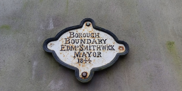 Schwarze Metalltafel an einem Gebäude mit weißer "Gemeindegrenze Edm Smithwick Bürgermeister"-Aufschrift in fetter Schrift.