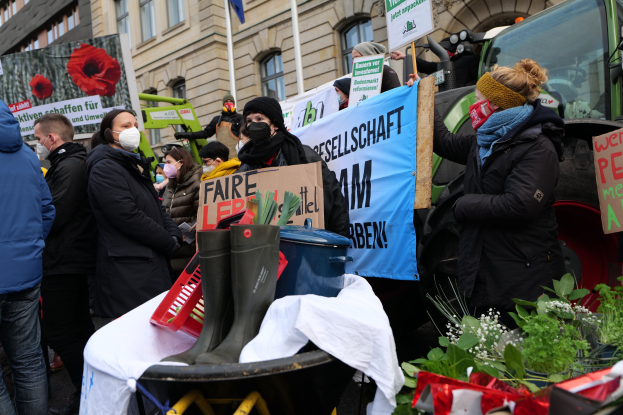 Eine Gruppe von Menschen mit Masken und Schildern protestiert vor einem Lastwagen, mit einem Tisch, Pflanzen und einer Flagge im Hintergrund.