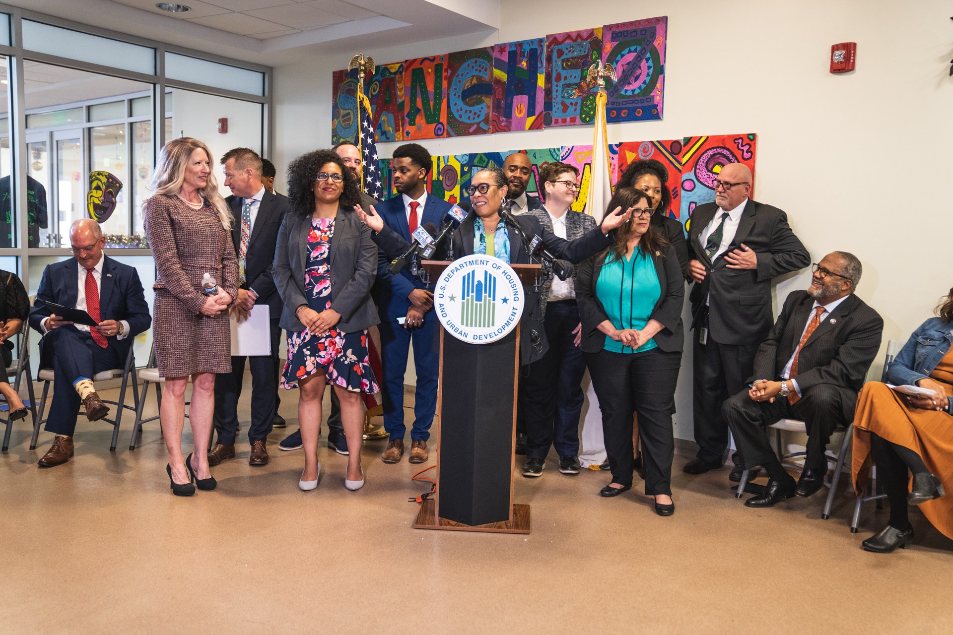 Gruppe von Menschen vor einem Rednerpult mit Mikrofon und einer Tafel mit der Aufschrift "Sancho Stadtrat tritt dem Stadtrat bei", mit sitzenden und stehenden G├Ąsten, Fahnen, Feuerl├Âscher und Deckenleuchten im Hintergrund.