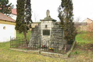 Ein steinernes Denkmal auf einer Rasenfläche, umgeben von einem Metallzaun und geschmückt mit Blumensträußen, mit Bäumen, Häusern und einer Wand im Hintergrund unter einem klaren blauen Himmel, als Holocaust-Gedenkstätte in Sibiu, Rumänien.