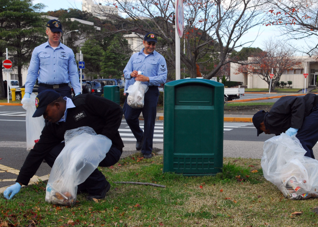Drei Polizeibeamte in Uniform sammeln Müll an einer Straße, mit Gebäuden, Fahrzeugen und Bäumen im Hintergrund.
