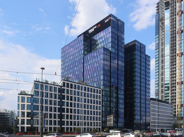 Großes Gebäude mit vielen Fenstern, identifiziert als Deutsche Bank Hauptquartier, in einer städtischen Umgebung mit Straßeninfrastruktur, Fahrzeugen, Grünflächen und einem bewölkten Himmel.