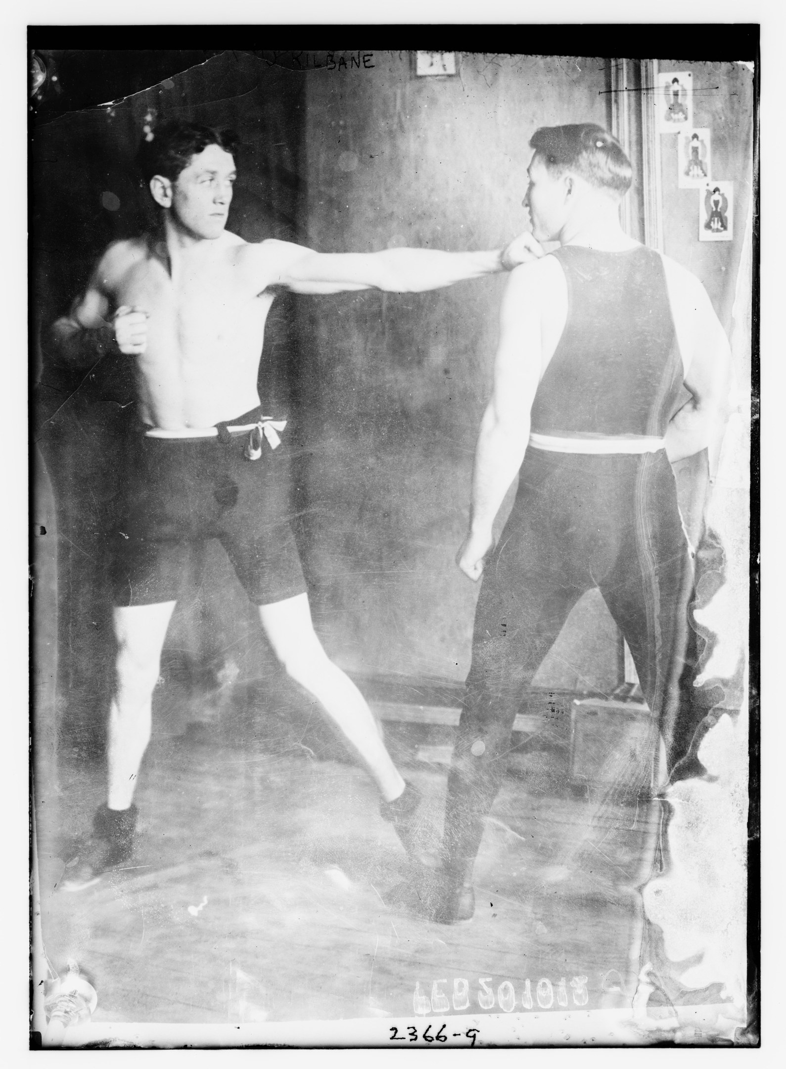 Schwarzes und weißes Foto von zwei Boxern in einem Ring mit Plakaten an der Hintergrundwand und Text unten.