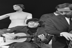 Ein Mann in einem schwarzen und weißen Outfit sitzt auf einem Stuhl und spielt eine Gitarre neben einer Schaufensterpuppe, mit einem Mikrofon vor ihnen, vor einem dunklen Hintergrund, als Tribut an den verstorbenen Paul McCartney.