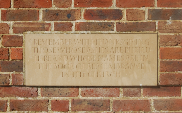Eine Backsteinmauer mit einer Tafel, auf der steht: "Erinnere dich mit Dankbarkeit an diejenigen, deren Asche hier begraben ist und deren Namen im Buch der Erinnerung in der Kirche stehen."