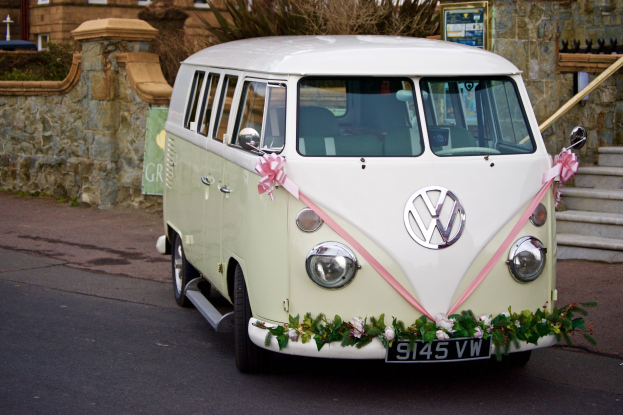 Weißer und rosafarbener VW Campervan, geschmückt mit Blumen und Blättern, vor einer Steinmauer mit Treppe auf der rechten Seite und einem Gebäude im Hintergrund geparkt.