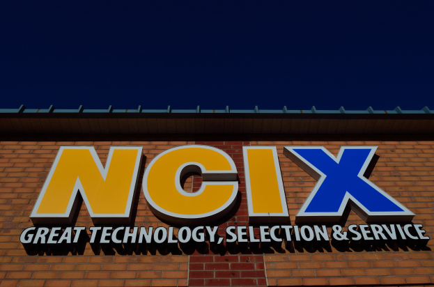 Außenansicht eines Backsteingebäudes mit einer Tafel, auf der "NCIX Great Technology, Selection & Service" steht, vor einem klaren blauen Himmel.
