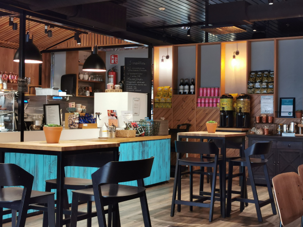 Innenraum eines Restaurants mit Holztischen, -stühlen, einer Topfpflanze auf einem Tisch, einer Schiefertafelmenü, einem Kühlschrank, einem Feuerlöscher, Regalen mit Flaschen, Schränken mit Gegenständen, einem Rahmenfoto an der Wand, Deckenleuchten und Lampen.