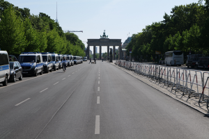Eine Reihe von Polizeiwagen auf einer Straße vor dem Brandenburger Tor in Berlin, Deutschland, mit Menschen auf Fahrrädern und Stehenden, Barrieren, Bäumen und einem Bogen mit Statuen im Hintergrund.