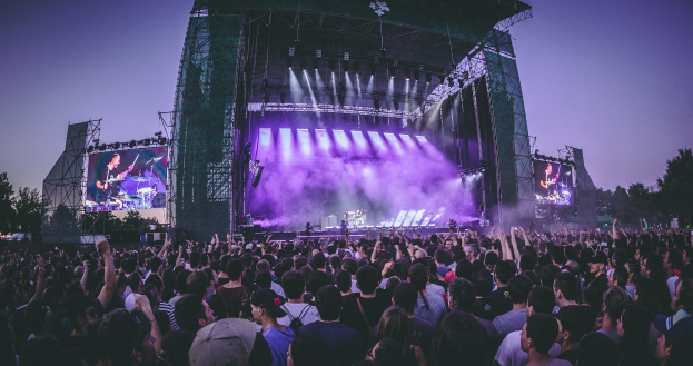 Eine große Menschenmenge auf einem Musikfestival schaut auf einer beleuchteten Bühne mit Instrumenten und Bildschirmen, die vor einem Gebäude, Bäumen und einem blauen Himmel aufgebaut sind.