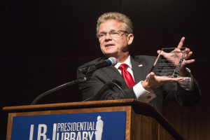 Ein Mann im Anzug und mit Krawatte, der eine Auszeichnung auf einem Podium mit Mikrofon und Tafel hält, vor einem dunklen Hintergrund bei einer Bibliothek-Veranstaltung.