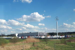 Ein Mercedes-Benz-Werk in Deutschland mit Fahrzeugen auf der Straße, umgeben von Gras, Pflanzen, Bäumen, Gebäuden und Masten, von denen einer ein Logo trägt, unter einem bewölkten Himmel.