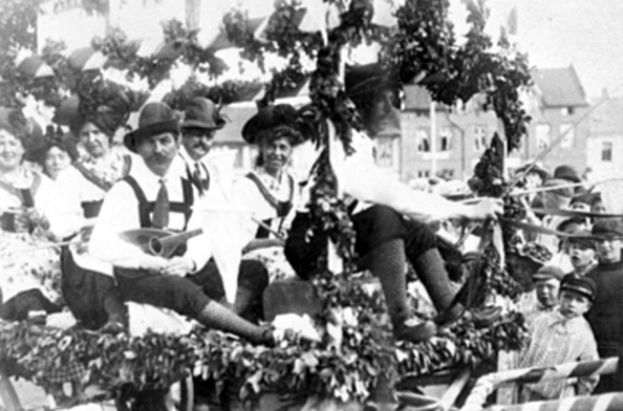 Schwarzes und weißes Bild eines Paradewagens mit Menschen in traditioneller bayrischer Tracht, einige spielen Instrumente, mit Blattdekorationen vor einem Hintergrund von Gebäuden unter einem klaren Himmel.