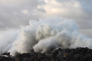 Eine große Welle kracht gegen die Felsen am Strand, mit Wasser, das gegen die Küste schwappt und einem bewölkten Himmel im Hintergrund.